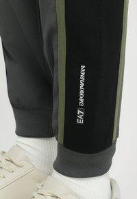 Pantalons de sport noirs avec des bandes latérales vertes, fabriqués en matériau lisse. Présentent un poignet élastique et une étiquette de logo blanche sur le côté.