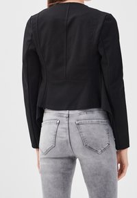 Veste noire légèrement courte à manches longues et col arrondi, associée à un jean en denim gris clair avec poches arrière et texture délavée.