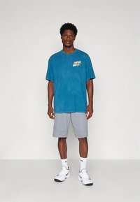 Türkiisivärvi üledimensioneeritud T-särk graafilise logoga rinnal, mida kannavad hallid spordipüksid ja valged tossud. Sujuv kangatekstuur.