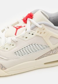 Zapatilla deportiva beige claro con detalles de malla, logo rojo y suela de goma texturizada. Cuenta con cordones blancos y un detalle sutil con patrón en el lateral.