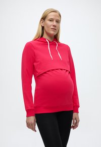 Raskaana oleva nainen, joka käyttää kirkkaanpunaista äitiysvetoketjullista hupparia, jossa on valkoiset nyöritykset, ja mustia leggingsejä, seisoo yksinkertaista vaaleaa taustaa vasten.