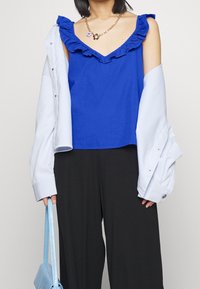 Haut bleu sans manches avec col à volants, associé à une chemise oversize bleu clair et un pantalon large noir. Tenant un sac bleu clair.