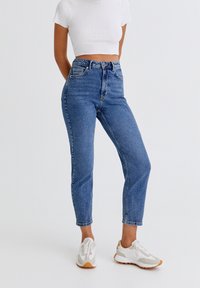 Jeans de cintura alta de color azul con una textura de denim clásica, que presentan un corte recto y un sutil desgastado, combinados con zapatillas blancas.