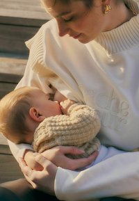 Crème kleurige sweatshirt met geribbelde kraag, met geborduurde tekst. Een kind in een textuurbeige gebreide trui is aan het borstvoeden.