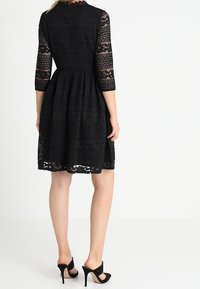 Svart spetsklänning med trekvartsärmar, intrikata mönster, en åtsittande midja och en utsvängd kjol. Bärs med svarta högklackade skor.