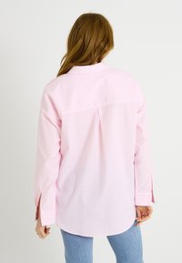 Camisa de rayas en rosa claro con mangas largas y cuello abotonado. Presenta un pliegue en la parte trasera y aberturas laterales en los puños. Textura de tela suave.