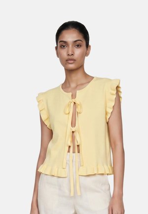 Woman wearing a sleeveless pale yellow top with front ties and ruffled edges paired with light beige pants.