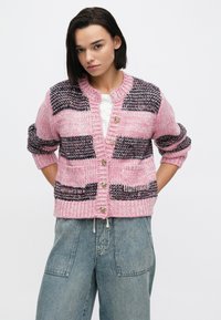 Cardigan roz tricotat cu modele alb-negru, având tiv cu dungi, decolteu rotund și nasturi aurii ca închidere. Mâneci cu croială lejeră.