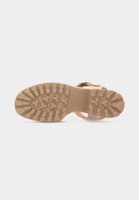 Σόλα από μπεζ σανδάλι με βαθιά τριγωνικά και ορθογώνια σχέδια πέλματος, ορατές αγκράφες ιμάντα και επίπεδη φτέρνα σε ανοιχτό φόντο.