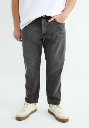 Osoba nosiaca vyblednuté čierne rovné rifle, biele tenisky s gumovou podrážkou a bielu košeľu, stojaca s jednou rukou v vrecku.