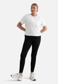 Vit bomull t-shirt i kombination med svarta ribbade leggings, bärs med vita sneakers. Modellen står med en fot framåt och händerna på höfterna.