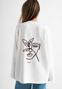 Witte sweatshirt met een bruin lijn-tekening gezicht en bladvorm ontwerp op de achterkant. Lange mouwen en een ontspannen pasvorm. Minimalistische esthetiek.
