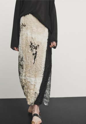 Personne portant une longue jupe froissée avec des motifs abstraits noirs et beiges, un haut noir à manches longues et des sandales noires avec des accents dorés.