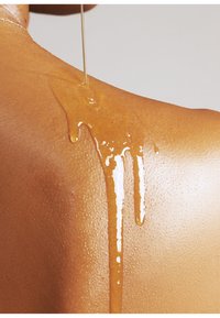 Guldolja som droppar ner på en texturerad brun hudytan, vilket skapar blanka höjdpunkter och släta reflexer mot en neutral bakgrund.