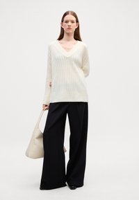 Sweter z dekoltem w serek w kolorze kremowym, wykonany z dzianiny o prążkowanej fakturze, w połączeniu z czarnymi spodniami o szerokich nogawkach i wysokim stanem. Trzyma jasnobeżową torebkę.