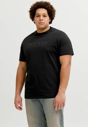 Mann med krøllete hår iført en svart "Originals" t-skjorte og lyseblå jeans, stående mot en enkel lys bakgrunn.