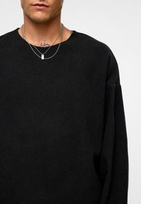 Zwarte crewneck trui met een gestructureerde afwerking. Heeft verlaagde schouders en gelaagde zilveren kettingen met een rechthoekige hanger.