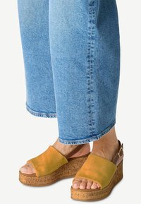 Keil-Sandalen mit einem Obermaterial aus gelbem Wildleder, einer korktexturierten Sohle und einem braunen Knöchelriemen. Getragen mit hellblauen weit geschnittenen Jeans.