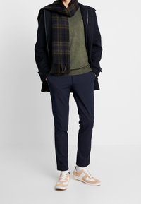 Marineblauer Mantel, olivgrüner Pullover, karierter Schal, dunkle Hose und beiges Sneakers mit weißen Akzenten. Strukturierte Materialien und modernes Design.