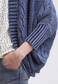 Cardigán de punto trenzado en azul profundo con patrones texturizados, que presenta secciones caladas y mangas a rayas, superpuesto a una blusa de lunares.