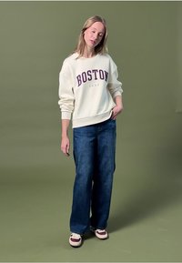 Krémová mikina se vzorem "BOSTON" vytištěným tmavě fialovou barvou, sladěná s volnými modrými džíny a vícero barvnými teniskami na zeleném pozadí.