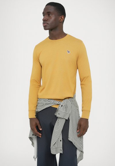 Maglietta a maniche lunghe colore giallo senape con un piccolo logo animale; abbinata a una camicia a quadri bianca e nera annodata in vita.