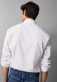 Man with dark hair wearing a white long-sleeve shirt with sleeves rolled up and blue jeans, facing a plain gray background.