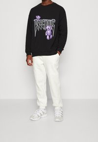 Uomo che indossa una felpa nera con orsetti di peluche viola e la scritta "PRECIOUS", pantaloni jogging bianchi con texture e sneakers bianche, in piedi con una mano in tasca.