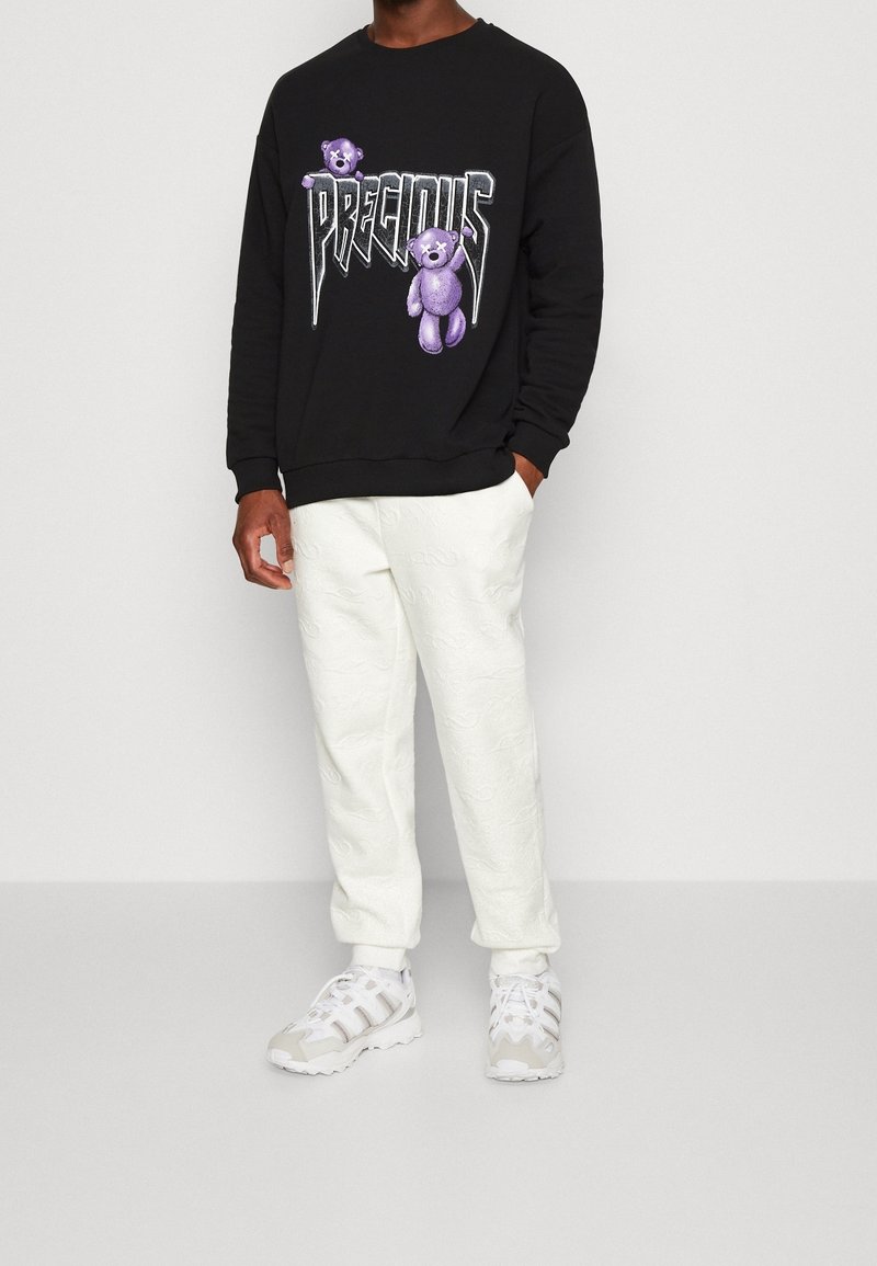 Uomo che indossa una felpa nera con orsetti di peluche viola e la scritta "PRECIOUS", pantaloni jogging bianchi con texture e sneakers bianche, in piedi con una mano in tasca.