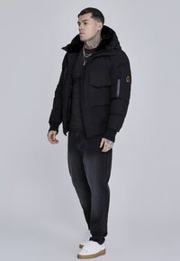 Black puffer jacket with a hood, featuring multiple front pockets, a textured fabric, and a logo patch on the arm. Worn over a dark top and jeans.
