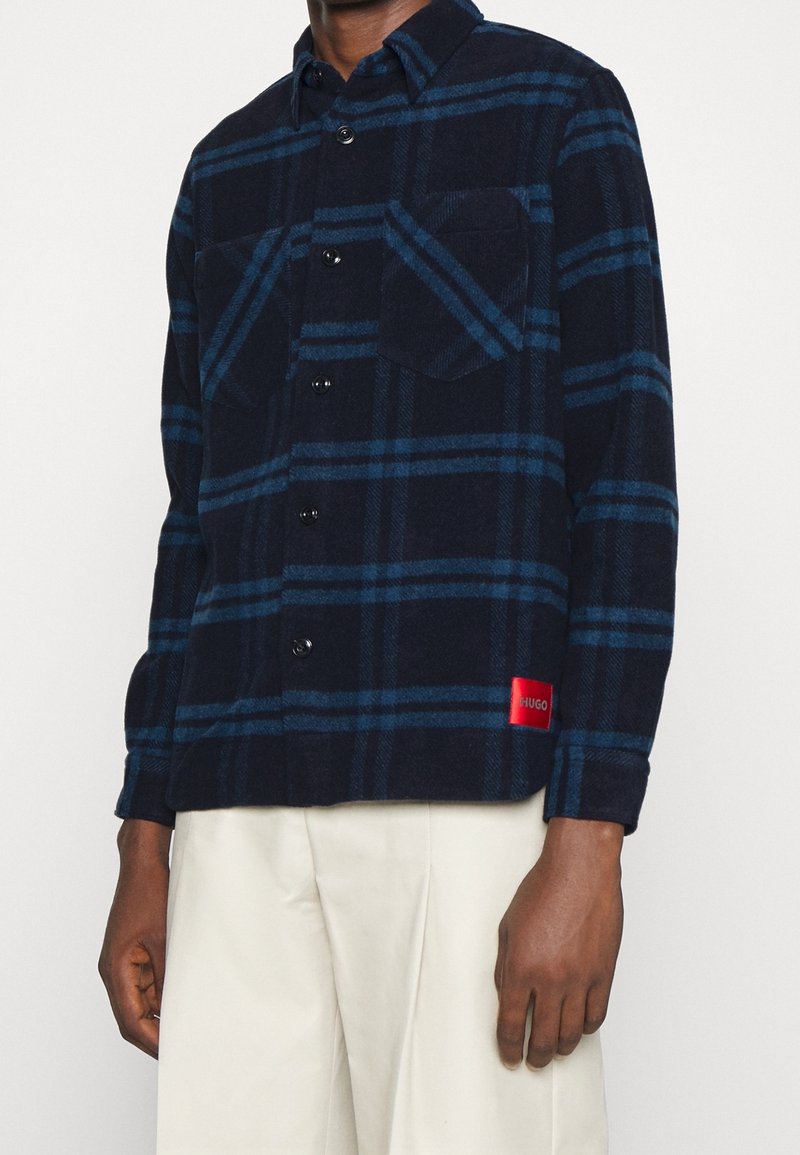 Homme portant une chemise à carreaux bleu marine et vert foncé avec poches poitrine et étiquette rouge HUGO, associée à un pantalon couleur crème.