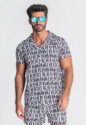Homme portant un ensemble assorti noir et blanc composé d'une chemise à manches courtes et d'un short avec un motif répétitif de texte gothique en gras, lunettes de soleil bleues réfléchissantes.