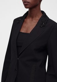 Femme portant un blazer noir avec un détail de col froncé et un top noir côtelé en dessous, montrée des épaules jusqu'au milieu du torse.
