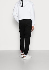 Man in een witte hoodie met zwart ALPHA-logo op de capuchon, zwarte cargo-broeken, witte sokken en witte sneakers met bruine hielaccenten.