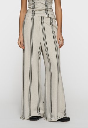 Donna che indossa pantaloni beige a gamba larga con strisce verticali nere e top senza maniche coordinato, in piedi con le mani rilassate ai lati.