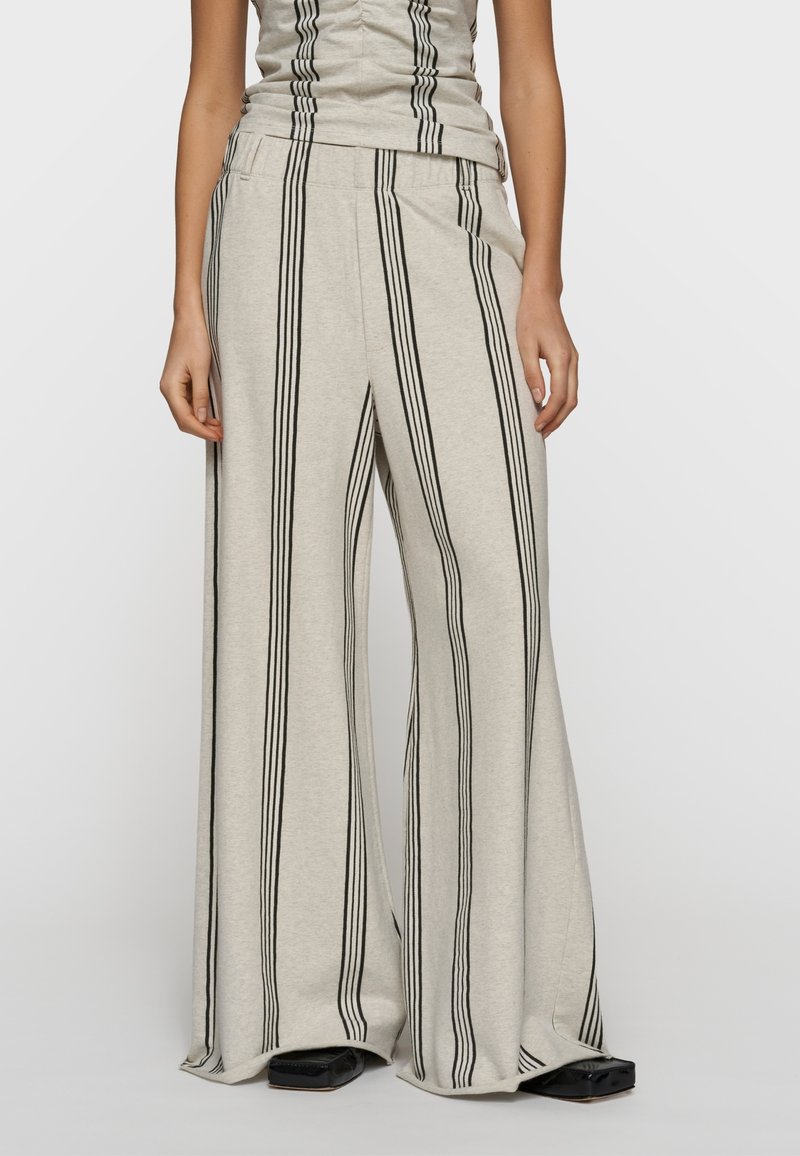 Kobieta w beżowych spodniach o szerokich nogawkach z pionowymi czarnymi paskami i dopasowanym topie bez rękawów, stojąca z rękami swobodnie opuszczonymi wzdłuż ciała.