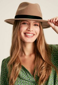 Brown felt fedora with a black band, featuring a wide brim, worn by a person in a green patterned blouse with long, wavy hair.