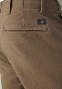 Brown cotton pants with a smooth texture, featuring a back pocket with a horizontal opening and a small navy logo label above it.