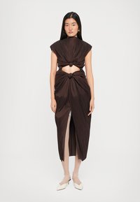 Robe brune plissée avec des détails en nœud au niveau du buste et de la taille, avec des manches courtes et une fente haute sur la cuisse. Design minimaliste, tissu texturé.