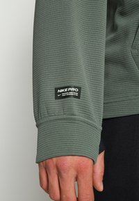 Gros plan sur une main portant un maillot à manches longues Nike Pro vert texturé avec une étiquette Nike Pro noire au poignet.