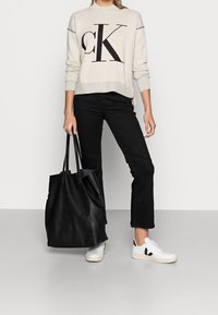 Personne portant un pull crème CK, un pantalon noir, des baskets blanches et noires, tenant un grand sac fourre-tout noir devant un fond uni.