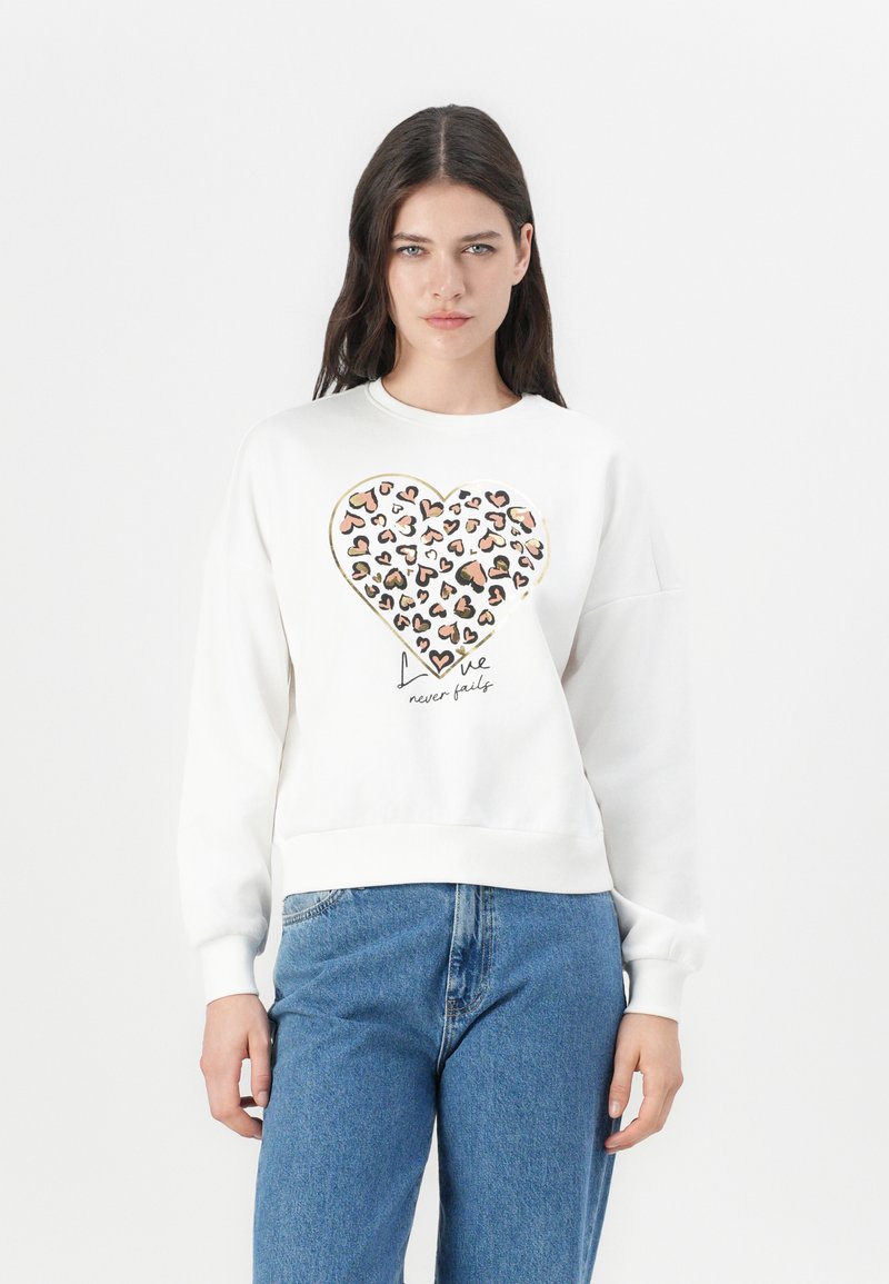 Weißer Sweatshirt mit einem großen Herzmotiv, das aus unterschiedlich gefärbten Herzen besteht, und dem Text "Liebe versagt nie" darunter. Bequemer Schnitt.