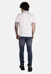 Polo bianco con bordi blu navy, jeans e sneakers nere. Modello mostrato di spalle con capelli ricci e tatuaggi visibili sulle braccia.