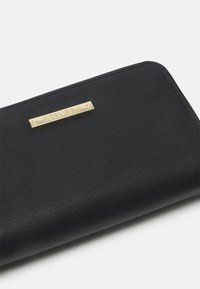Portefeuille noir avec une surface en tissu texturé, avec une plaque de logo dorée mise en avant sur le devant. Forme rectangulaire, fermeture à glissière.