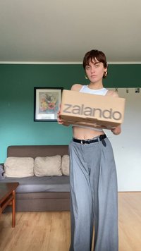 Kartonasti paket označen z "zalando," drži se pred nevtralno ozadje. Model nosi kratko belo majico in siva široka hlače.