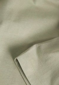 Hellgrüner Baumwollstoff mit glatter Textur. Zeigt eine gefaltete Kante und sichtbare Nähdetails am Saum.