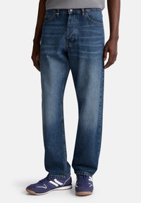 Tmavě modré džíny s uvolněným střihem, rovnými nohavicemi a viditelným švem. Nosí se s fialovými sportovními botami s bílými akcenty.