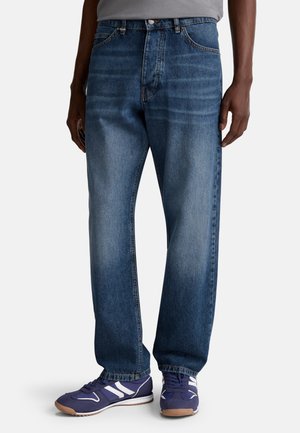 Donkerblauwe denim jeans met een relaxte fit, rechte benen en zichtbare stiksels. Draagt met paarse sportschoenen met witte accenten.