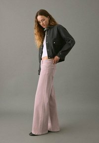 Chaqueta de mezclilla negra sobre una blusa blanca, combinada con pantalones de pierna ancha en color rosa pastel. Tejido suave y detalles mínimos, sobre un fondo gris.