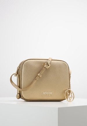 Sac bandoulière Armani Jeans en cuir doré texturé avec bandoulière réglable et breloque logo circulaire, posé sur une surface blanche.